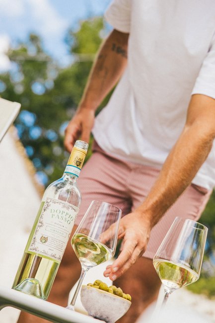 In una giornata di sole, un uomo versa con cura del vino bianco da una bottiglia con l'etichetta “Doppio Passo” accanto a una ciotola di olive.