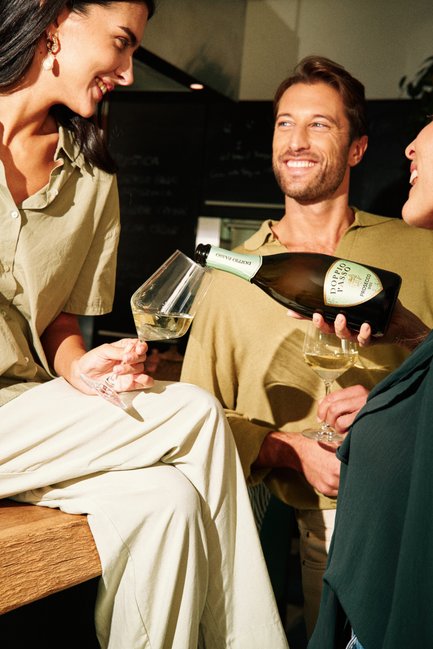 Una donna vestita di verde chiaro sorride mentre un uomo le versa del Prosecco nel bicchiere, circondata dagli amici in un ambiente accogliente.