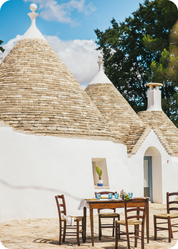 Charming trulli houses with conical roofs stand against a bright blue sky, featuring a rustic wooden table set for a meal.