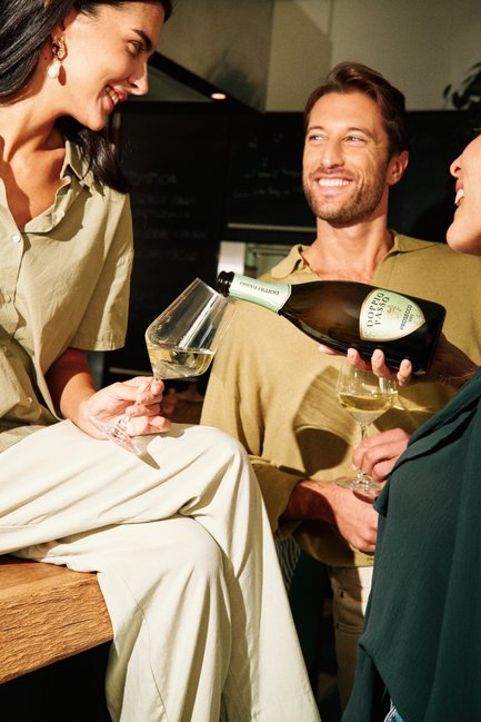 A woman in a light green outfit smiles as a man pours Prosecco into her glass, surrounded by friends in a cozy setting.