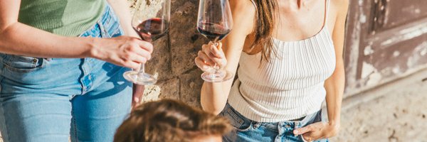 Drei Freunde genießen einen sonnigen Nachmittag, lachen und stoßen mit einem Glas Rotwein vor einer rustikalen Steinmauer an.