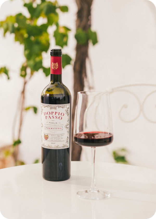 A bottle of Doppio Passo Primitivo wine stands next to a filled glass on a white table, surrounded by green vines.