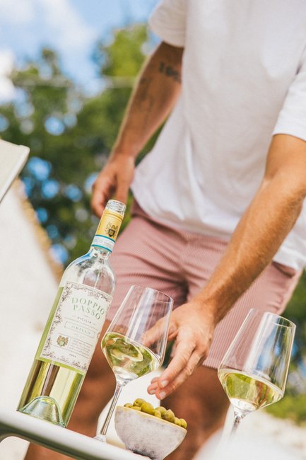 An einem sonnigen Tag schenkt ein Mann sorgfältig Weißwein aus einer Flasche mit der Aufschrift „Doppio Passo” neben einer Schale mit Oliven ein.