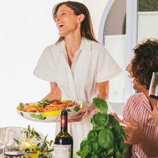 A joyful woman in a white dress serves a colorful salad to friends at a beautifully set outdoor dining table.