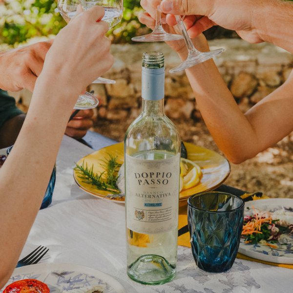 Eine Gruppe von Freunden stößt fröhlich mit Gläsern an einem sonnigen Tisch im Freien an, auf dem eine Flasche Doppio Passo-Wein steht.