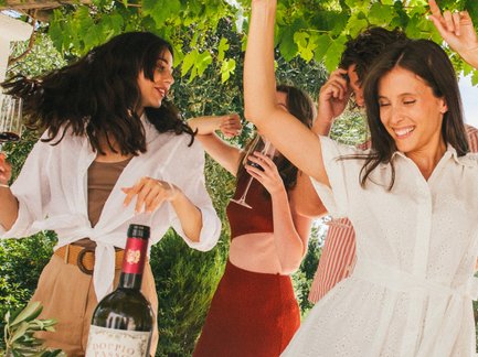 In einer lebhaften Atmosphäre tanzen Frauen in stilvollen Outfits, stoßen mit Wein an und genießen ein festliches Mahl unter einem grünen Blätterdach.