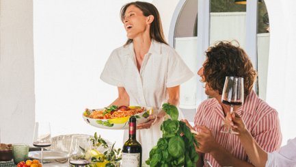 A joyful woman in a white dress serves a colorful salad to friends at a beautifully set outdoor dining table.