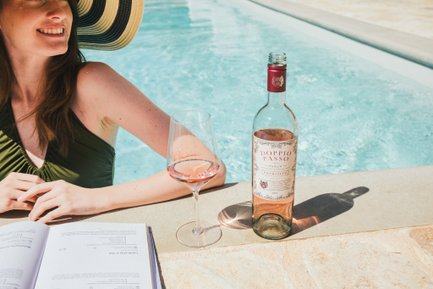 Una donna si rilassa a bordo piscina, gustando un bicchiere di vino rosato mentre legge un libro sotto il sole, indossando un elegante cappello da sole.