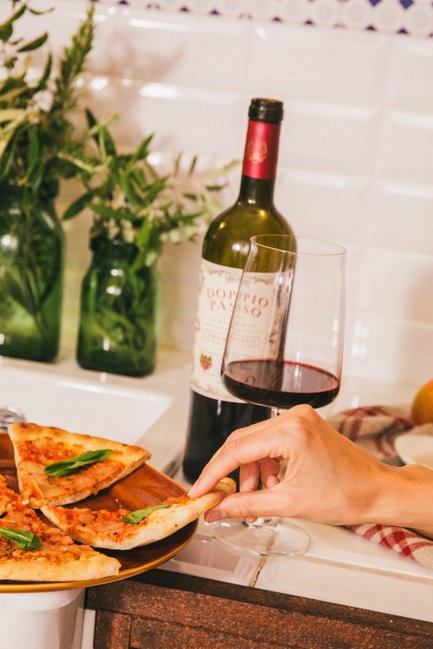 Leckere Pizzastücke mit frischem Basilikum werden auf einem Teller serviert, während eine Hand sich bereit macht, eines zu nehmen, und daneben steht ein Glas Wein.