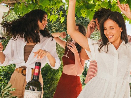 In un'atmosfera vivace, donne in abiti eleganti ballano e brindano con il vino, godendosi un pasto festivo sotto un baldacchino di foglie.