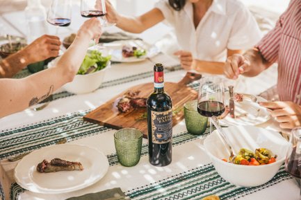 Hands reach for glasses of red wine in a sunlit gathering, featuring a variety of dishes on a beautifully arranged table.